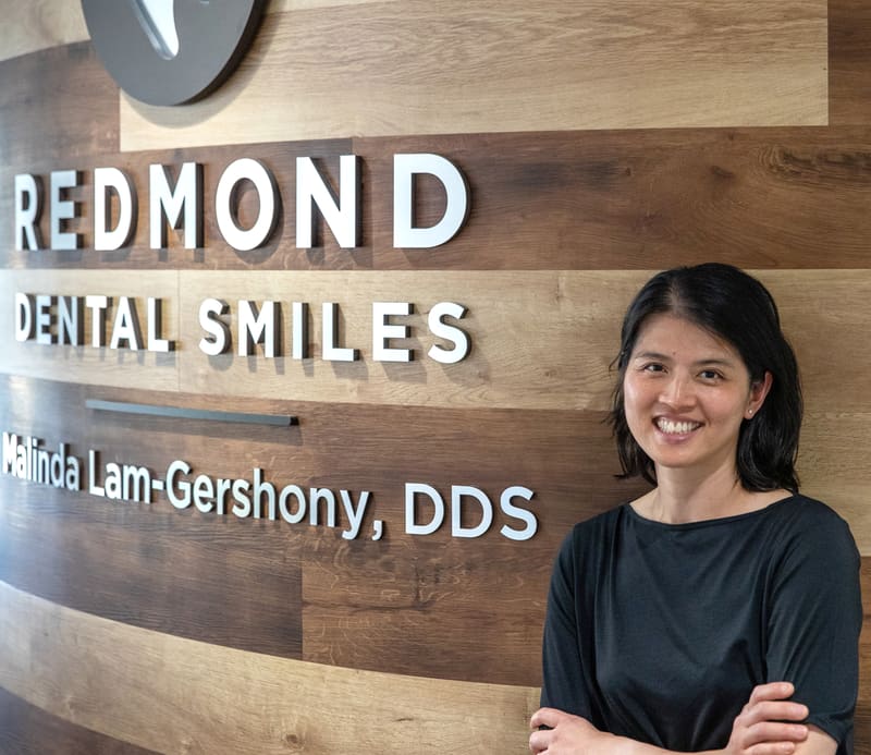 Dr. Malinda Lam-Gershony standing in front of the Redmond Dental Smiles logo wall