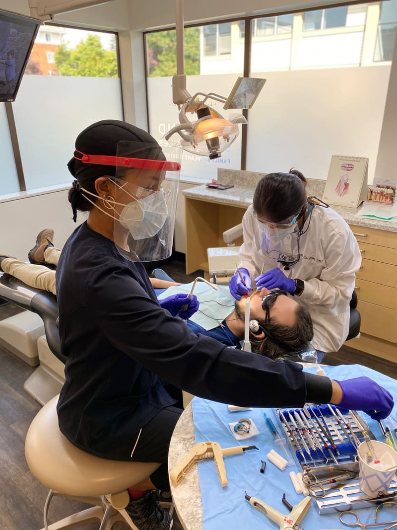 Dentist and assistant in face shields treating a patient in a bright treatment room